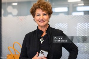 A poised middle-aged woman with curly hair and a stylish black jacket stands confidently in a modern office, holding her smartphone and glasses. Her warm yet professional expression conveys leadership, digital connectivity, and executive presence.
